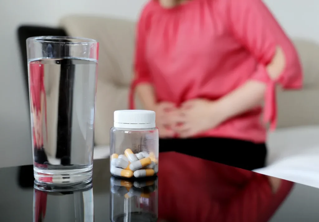 A woman sitting on a bed holding her stomach in discomfort, with medication on a table nearby, illustrating the connection between medicine and diarrhea.