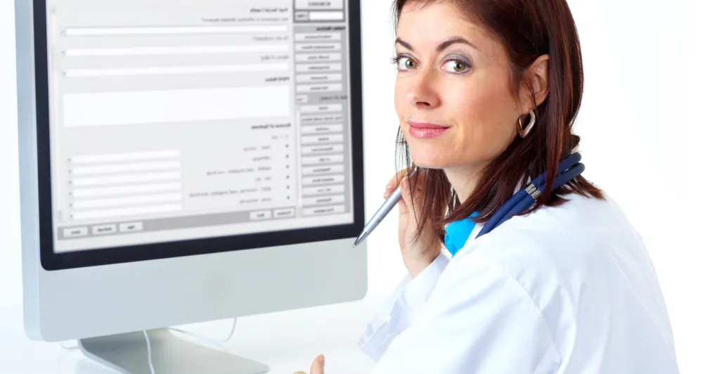 A doctor sits at her computer and looks over her shoulder at the camera.